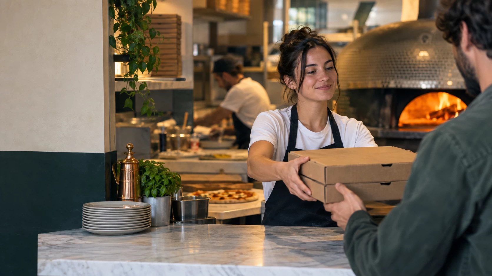 Addetta di una pizzeria consegna due scatole da asporto a un cliente al banco.
